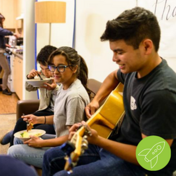 4 students interacting with each other, eating food, and playing guitar